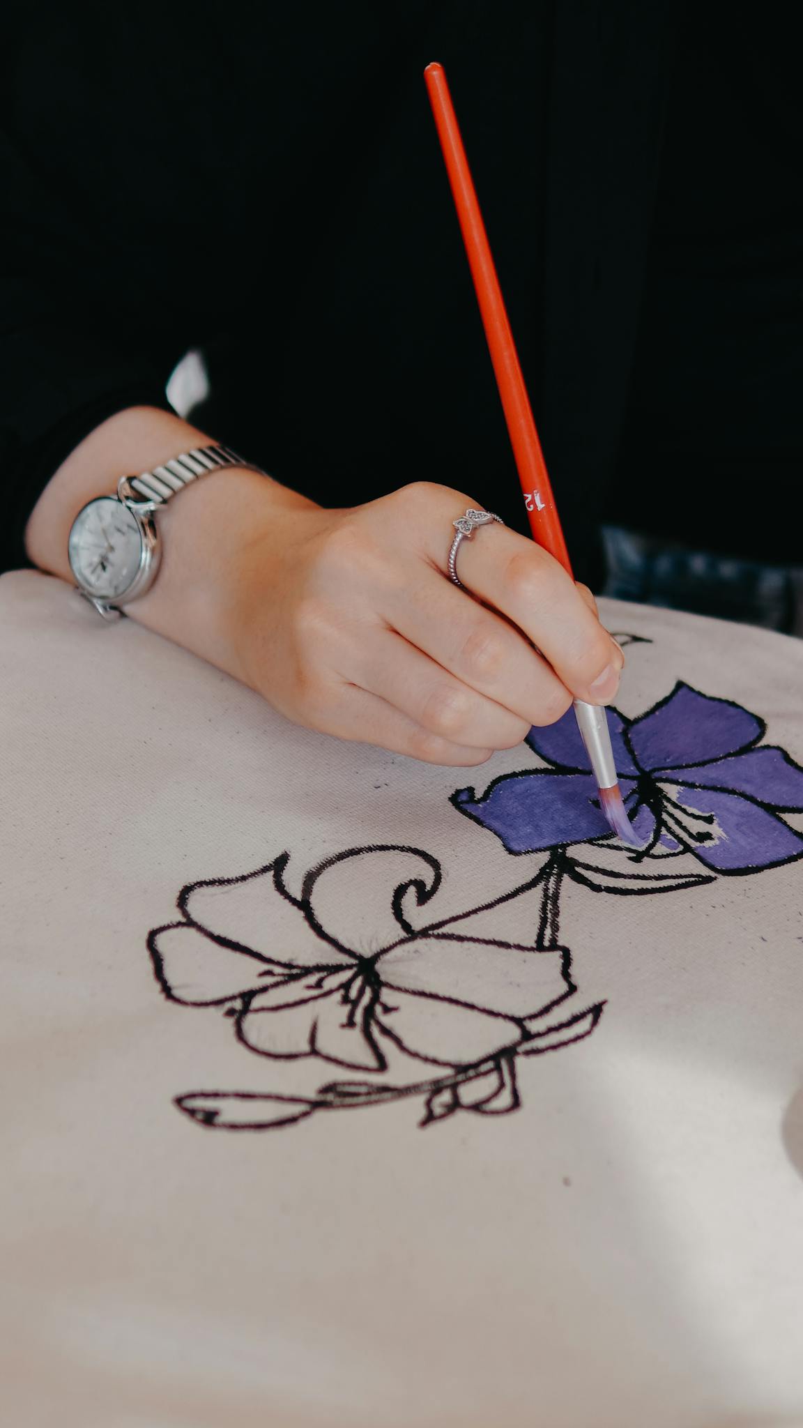Hand paints intricate floral design on fabric with red-handled brush, showcasing creativity.